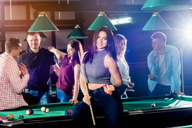 Bilardo oynayan genç bir kadın. Elinde ıstakayla masanın yanında poz veriyordu. Arka planda bir grup arkadaş.