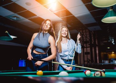 Bilardo oynayan iki genç kadın. Arka planda bilardo odası..