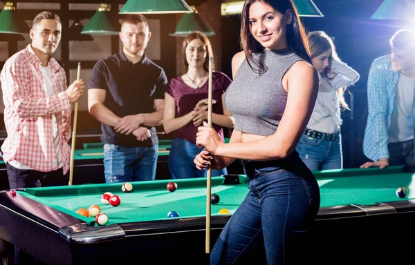 Young woman playing in billiard. Posing near the table with a cue in her hands. A group of friends on the background.