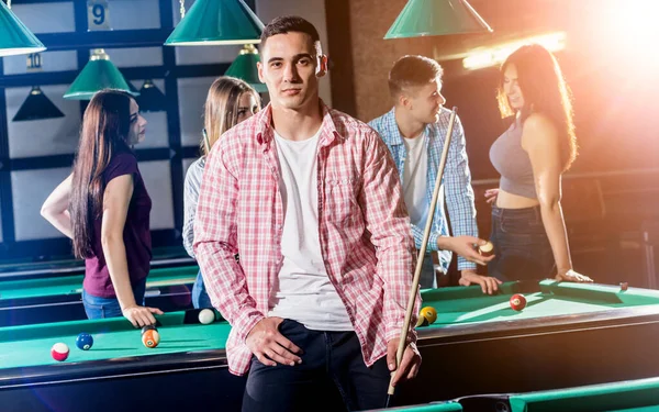 Young man playing in billiard. Posing near the table with a cue in his hands. His friends on the background.