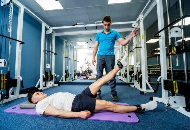 Rehabilitasyon terapisi. Fizyoterapistin gözetiminde paspas üzerinde egzersiz yapan genç bir adam. Omurgada tedavi ağrısı