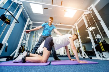 Rehabilitasyon terapisi. Fizyoterapistin gözetiminde paspas üzerinde egzersiz yapan genç bir adam. Omurgada tedavi ağrısı