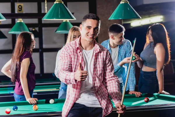 Young man playing in billiard. Posing near the table with a cue in his hands. His friends on the background.