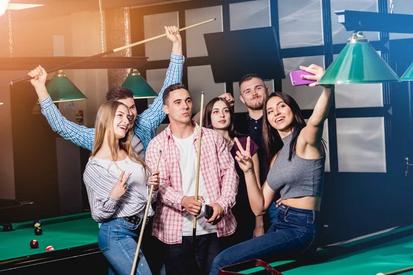 Un grupo de amigos hace una selfie en la mesa de billar. Posando con un taco en sus manos . — Foto de Stock