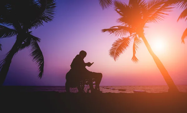 Disabled man in a wheelchair on the beach. Silhouette at sunset. — Stockfoto
