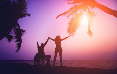Disabled man in a wheelchair with his wife on the beach. Silhouettes at sunset