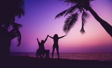 Disabled man in a wheelchair with his wife on the beach. Silhouettes at sunset