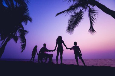 Disabled man in a wheelchair with his family on the beach. Silhouettes at sunset