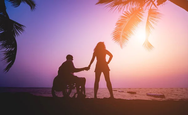 Um homem deficiente numa cadeira de rodas com a mulher na praia. Silhuetas ao pôr-do-sol — Fotografia de Stock