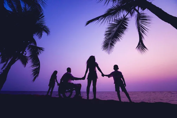 Disabled man in a wheelchair with his family on the beach. Silhouettes at sunset — Stok fotoğraf