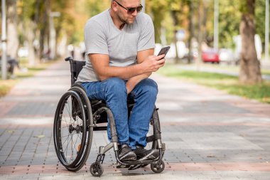 Park sokağındaki tekerlekli sandalyedeki engelli adam akıllı telefon kullanıyor.