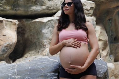 Hamile bir kadın zarif bir şekilde güzel bir şelale kayasının önünde duruyor..