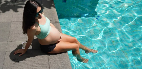 Turkuaz bikinili hamile bir kadın havuz kenarında oturuyor..