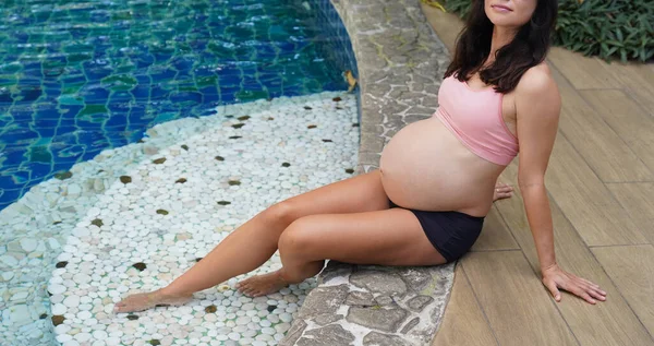 Pembe bikinili hamile bir kadın havuzun kenarında oturuyor..