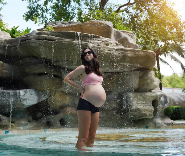 Hamile bir kadın zarif bir şekilde güzel bir şelale kayasının önünde duruyor..