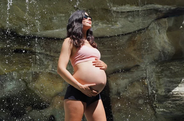 Hamile bir kadın zarif bir şekilde güzel bir şelale kayasının önünde duruyor..