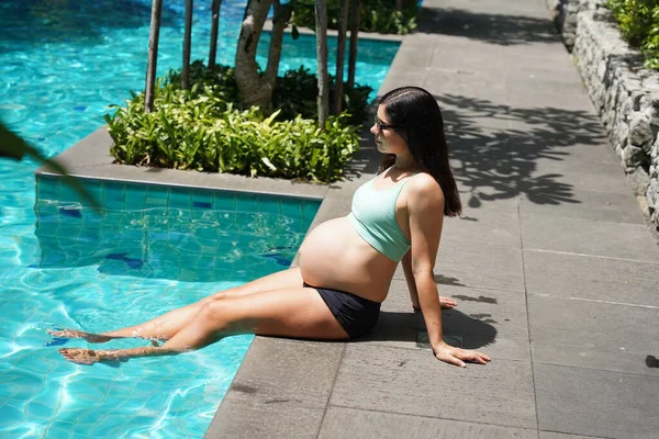 Turkuaz bikinili hamile bir kadın havuz kenarında oturuyor..