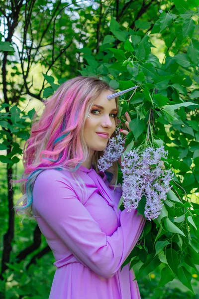 Fairy girl unicorn with rainbow hair and shiny makeup holding lilac flowers.