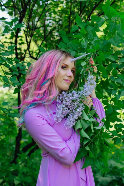 Fairy girl unicorn with rainbow hair and shiny makeup holding lilac flowers.