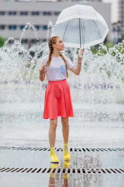 Genç güzel kız sarı çizmeli iki örgüler ve şeffaf şemsiye ile Çeşme duruyor. Şehirde yağmurlu gün.