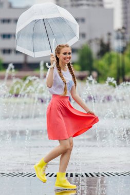 Genç güzel kız sarı çizmeli iki örgüler ve şeffaf şemsiye ile Çeşme duruyor. Şehirde yağmurlu gün.