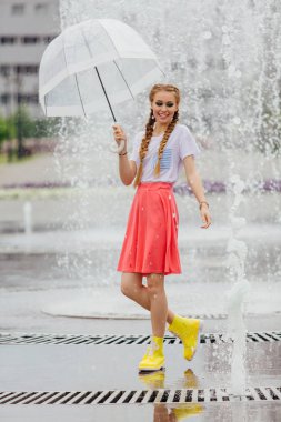 Genç güzel kız sarı çizmeli iki örgüler ve şeffaf şemsiye ile Çeşme duruyor. Şehirde yağmurlu gün.