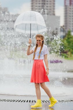 Genç güzel kız sarı çizmeli iki örgüler ve şeffaf şemsiye ile Çeşme duruyor. Şehirde yağmurlu gün.