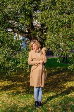 Güzel zarif sarışın kadın ceket ayakta sonbahar park ağaç altında giyinmiş. Güneşli Ekim gün.