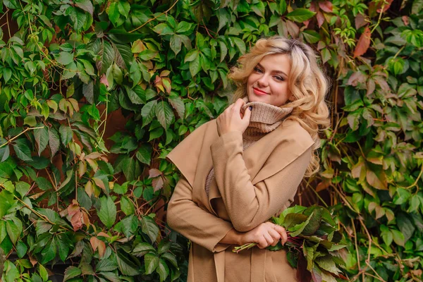 Yuong mooie vrouw in de trui en jas staat naast de achtergrond van wilde druiven houden boeket van de herfst bladeren — Stockfoto