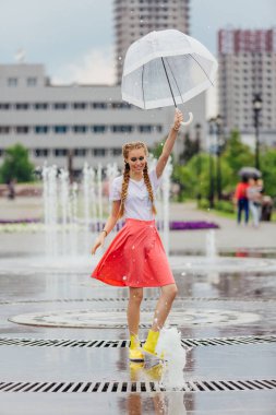 Genç güzel kız sarı çizmeli iki örgüler ve şeffaf şemsiye ile Çeşme duruyor. Şehirde yağmurlu gün.