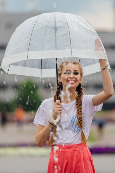 透明傘と黄色のブーツでお下げ髪若いきれいな女の子は噴水のそばに立ちます 雨の日の市 — ストック写真