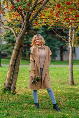 Güzel zarif sarışın kadın ceket ayakta rowanberries ve yaprakları oluşan sonbahar park holding üvez ağacının altında giyinmiş. Güneşli Ekim gün.