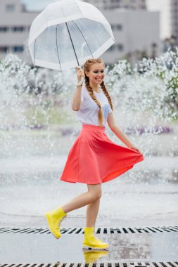 Genç güzel kız sarı çizmeli iki örgüler ve şeffaf şemsiye ile Çeşme duruyor. Şehirde yağmurlu gün.