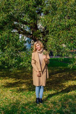 Güzel zarif sarışın kadın ceket ayakta sonbahar park ağaç altında giyinmiş. Güneşli Ekim gün.