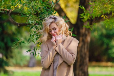 Güzel zarif sarışın kadın ceket ayakta sonbahar park ağaç altında giyinmiş. Güneşli Ekim gün.