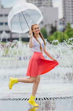 Genç güzel kız sarı çizmeli iki örgüler ve şeffaf şemsiye ile Çeşme duruyor. Şehirde yağmurlu gün.