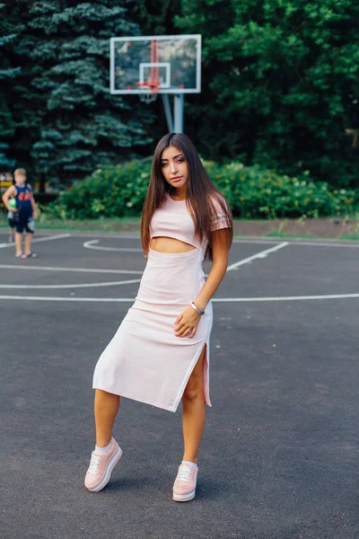 Charming brunette female dressed in a pink dress posing on the basketball court during sunset.