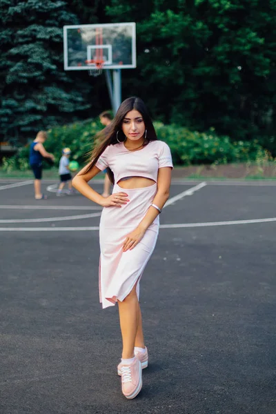 Charming brunette female dressed in a pink dress posing on the basketball court during sunset.