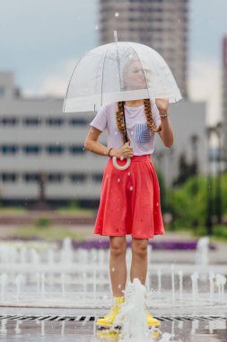 Genç güzel kız sarı çizmeli iki örgüler ve şeffaf şemsiye ile Çeşme duruyor. Şehirde yağmurlu gün.