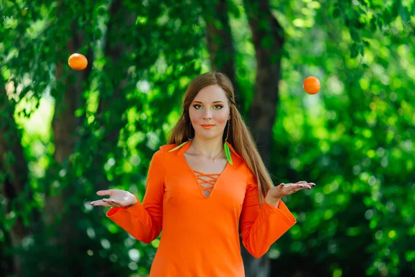 Portrait of pretty red hair woman throwing up two juicy delicious mandarins at summer green park.