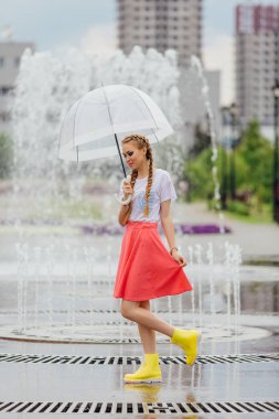 Genç güzel kız sarı çizmeli iki örgüler ve şeffaf şemsiye ile Çeşme duruyor. Şehirde yağmurlu gün.