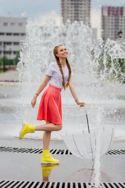 Genç güzel kız sarı çizmeli iki örgüler ve şeffaf şemsiye ile Çeşme duruyor. Şehirde yağmurlu gün.