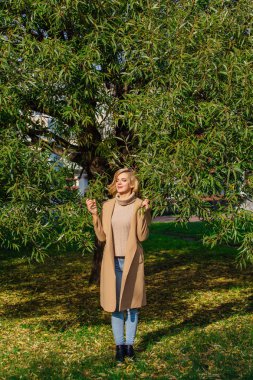 Güzel zarif sarışın kadın ceket ayakta sonbahar park ağaç altında giyinmiş. Güneşli Ekim gün.