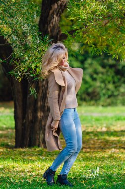 Güzel zarif sarışın kadın ceket ayakta sonbahar park ağaç altında giyinmiş. Güneşli Ekim gün.