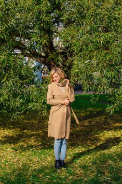 Güzel zarif sarışın kadın ceket ayakta sonbahar park ağaç altında giyinmiş. Güneşli Ekim gün.