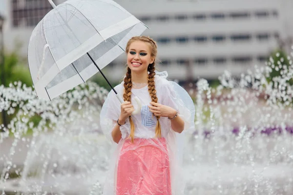 透明傘と黄色のブーツでお下げ髪若いきれいな女の子は噴水のそばに立ちます 雨の日の市 — ストック写真