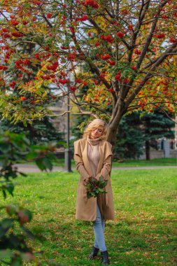 Güzel zarif sarışın kadın ceket ayakta rowanberries ve yaprakları oluşan sonbahar park holding üvez ağacının altında giyinmiş. Güneşli Ekim gün.
