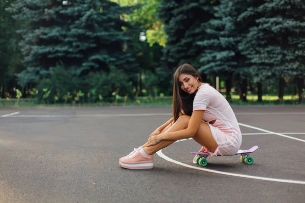 一个微笑迷人的黑发女子坐在篮球场的滑板上的画像 日落时分 穿着时髦的快乐女人正在休息 — 图库照片