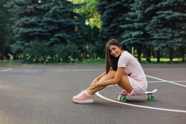 Portrait of a smiling charming brunette female sitting on her skateboard on a basketball court. Happy woman with trendy look taking break during sunset.