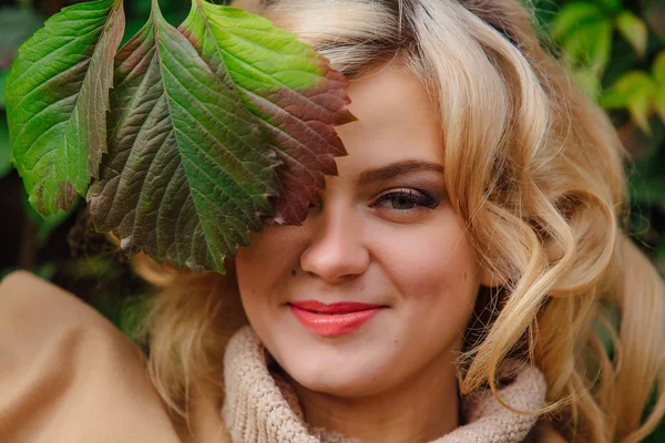 Young beautiful woman in sweater and coat stands next to the background of wild grapes holding leaves close to the face in autumn park